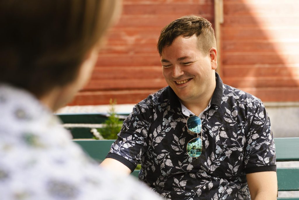 Smiling man wearing a dark floral pattern shirt with sunglasses hanging from his chest outdoors near wooden steps