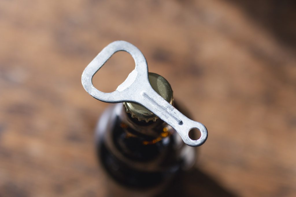 Top-down view of a metal bottle opener lifting a bottle cap off a dark glass bottle, with a warm brown background.