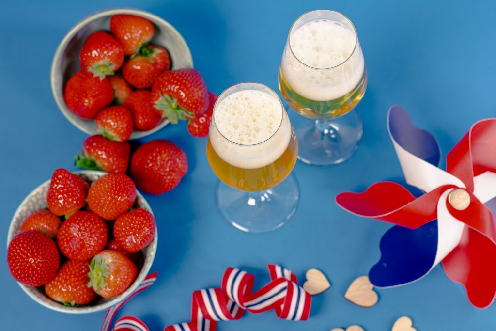 17maisiderciderfrokostoelakademiet202437 | Ølakademiet AS Bowl of strawberries with two frothy beer glasses on a blue table surrounded by redwhiteblue ribbons and wooden hearts for a festive scene