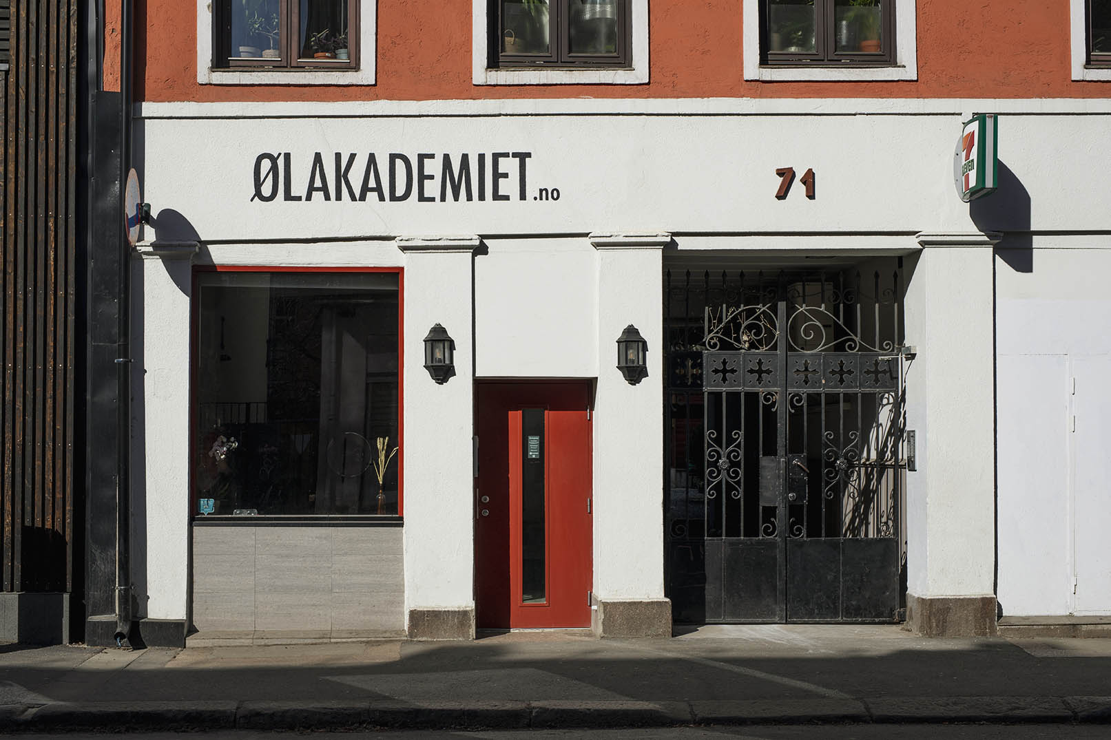 Fasaden | Ølakademiet AS White storefront with an orange upper façade a red central door and black wrought iron gate sign reads ØLAKADEMIETno 71 on the wall