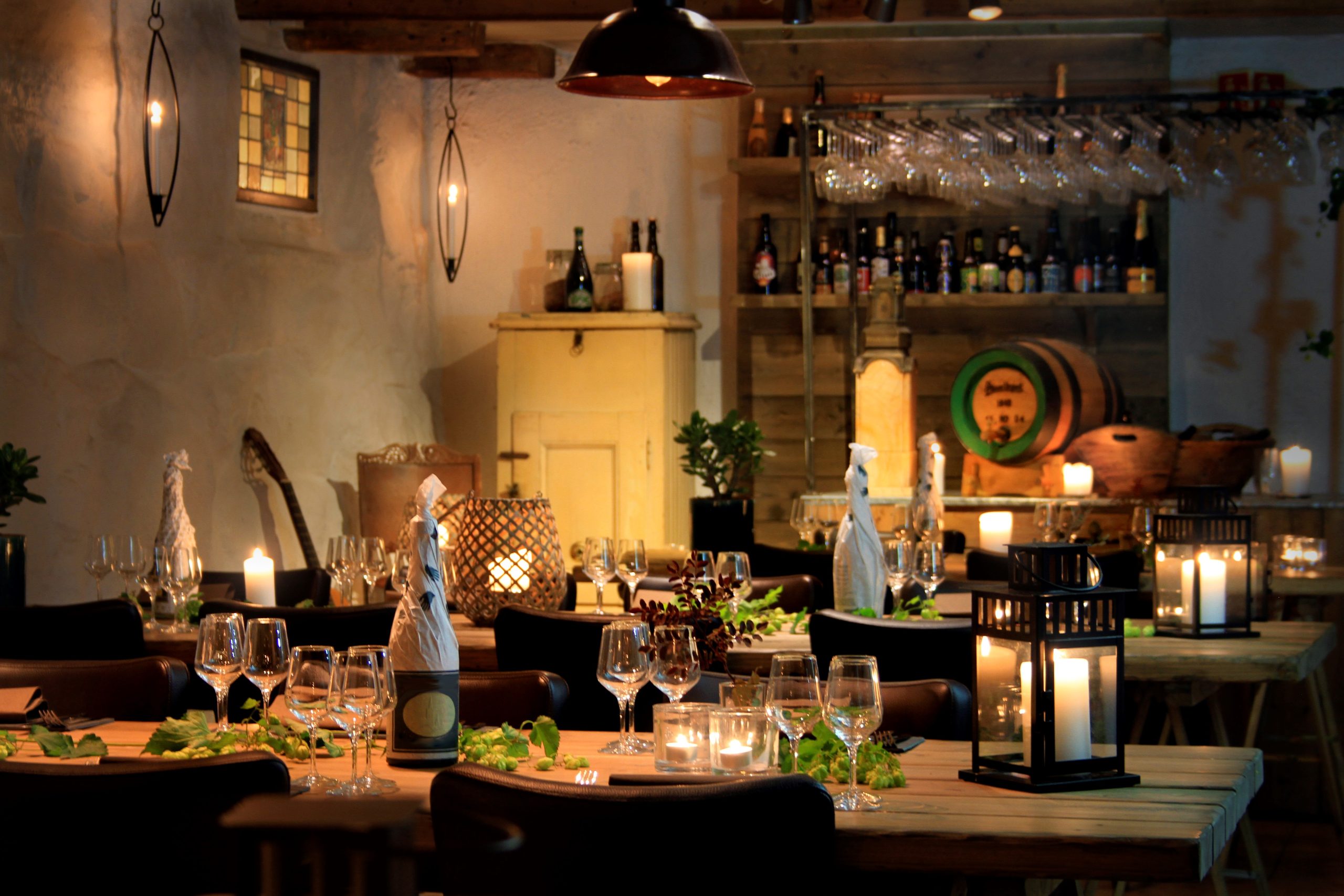 Dimly lit restaurant dining area with candlelit tables, wine glasses, and a back bar stocked with bottles.