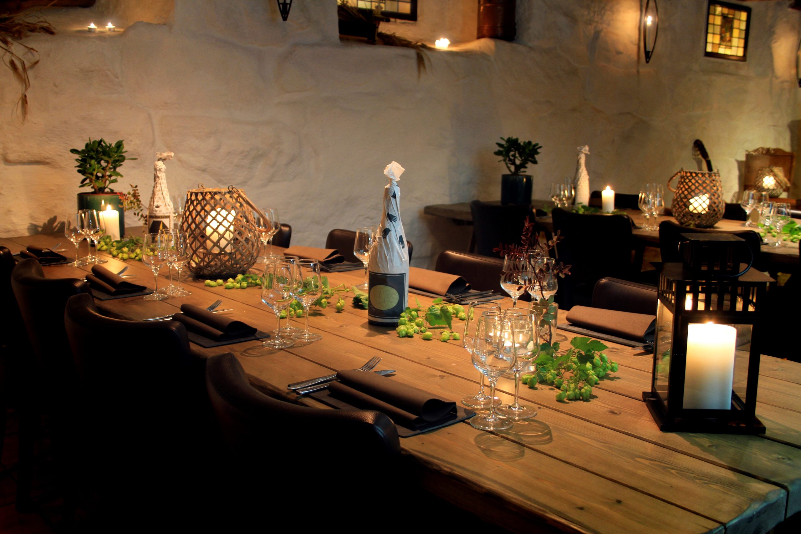 Long wooden dining table set for dinner with wine glasses, napkins, greenery, and warm candlelight in a rustic room.