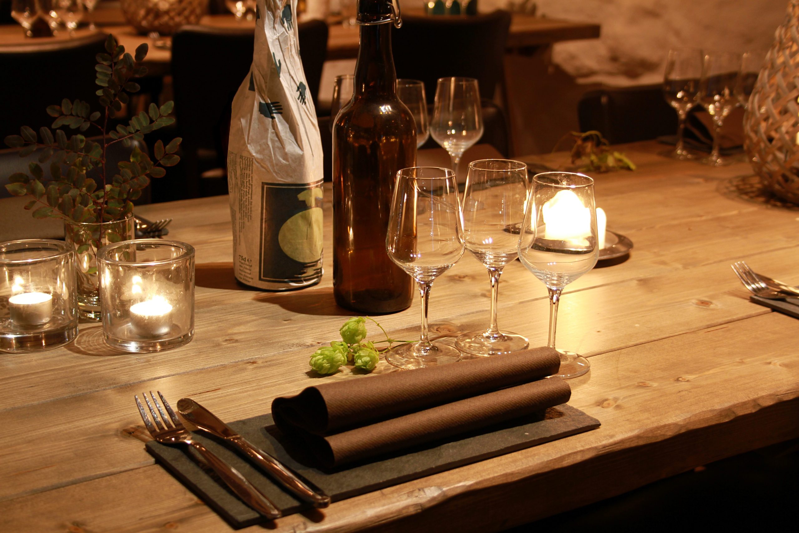 Rustic dining table set with a central wine bottle, candles, stemware, and greenery in warm light.