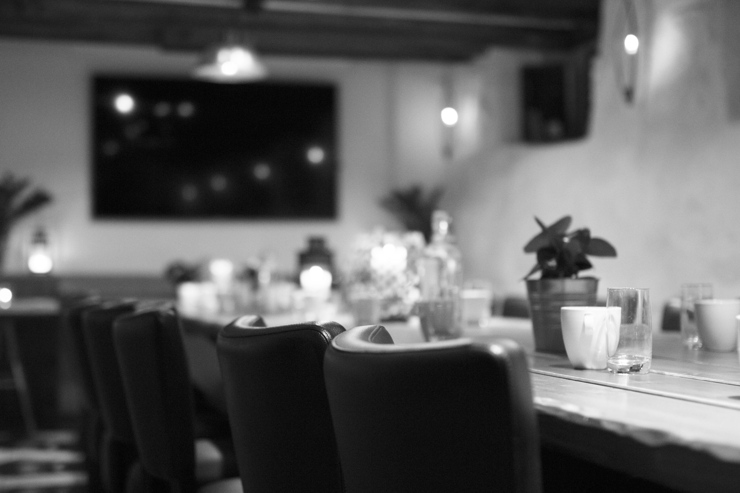 Dimly lit restaurant interior with table settings, glassware, and a potted plant in the foreground.