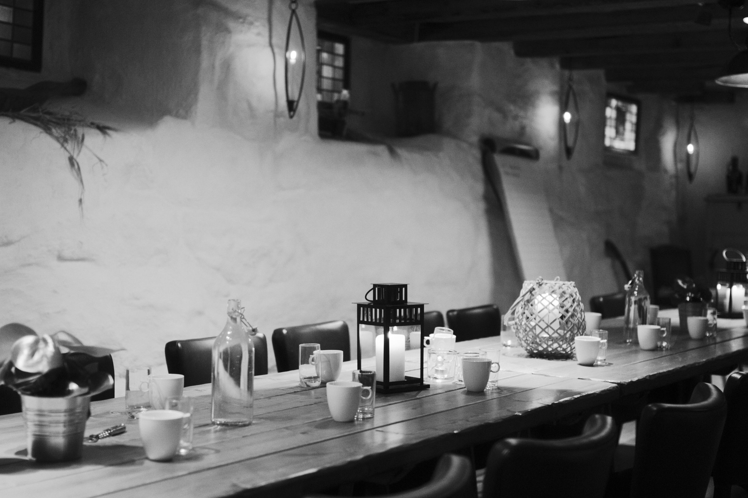 Long wooden communal table set for a meal with mugs, glasses, and lantern centerpieces in a dimly lit, cozy room.