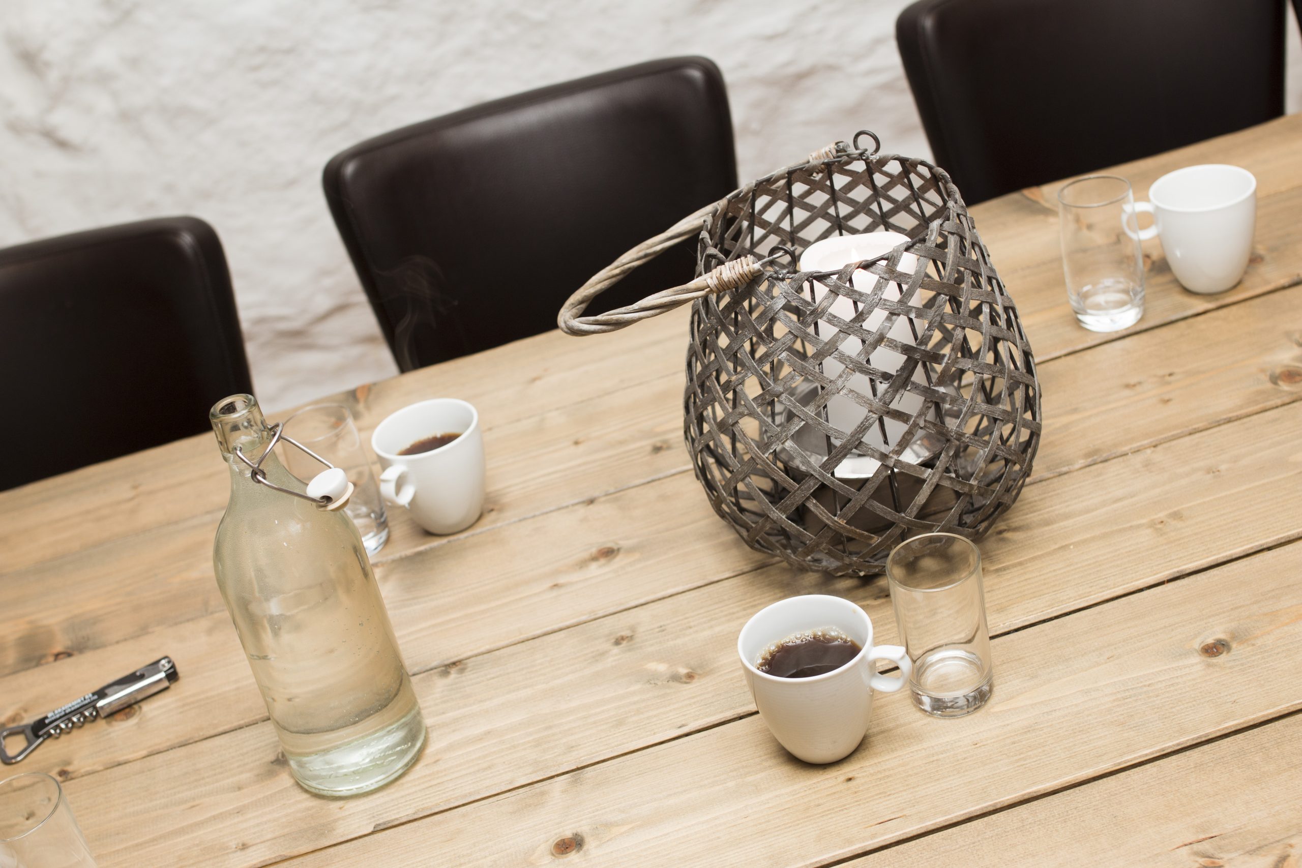 Wooden table with white mugs, glasses, a bottle, and a decorative metal lantern centerpiece in a casual setting.
