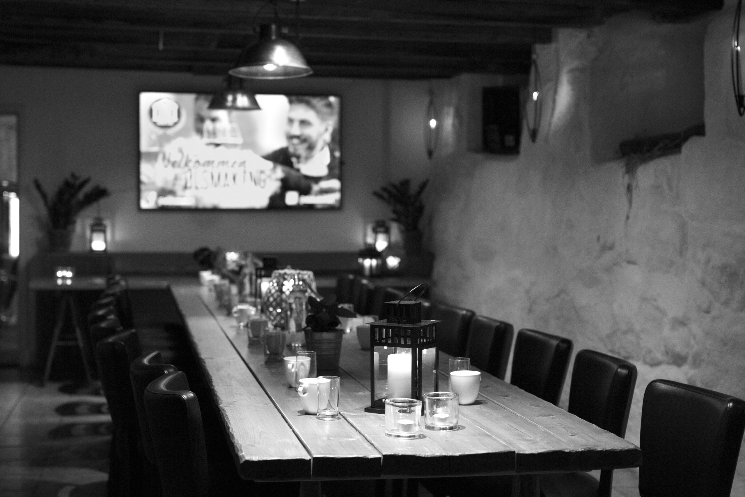 Dimly lit restaurant with a long wooden table set with mugs, candles, and small lanterns; blurred screen on the back wall.