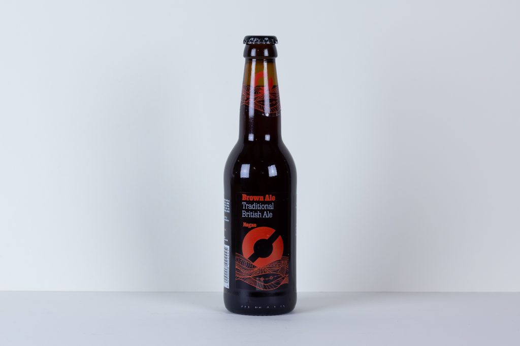 Brown beer bottle standing upright on a white surface with a red and black decorative label and cap in the center of the frame