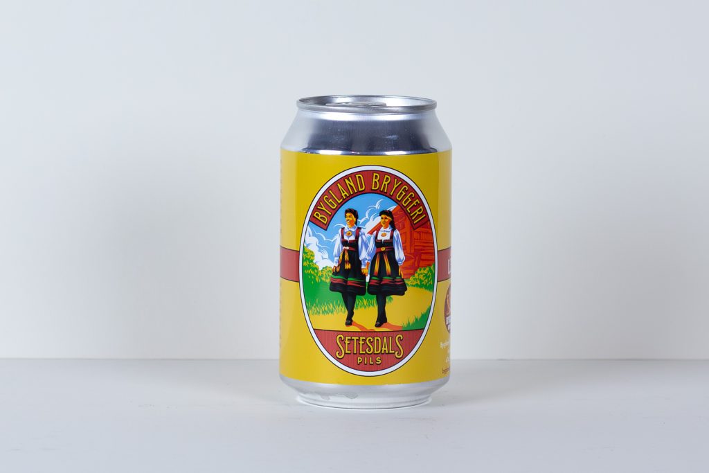 Yellow beer can with a colorful label featuring two women in traditional folk dress reads Bygland Bryggeri and Setesdals Pils