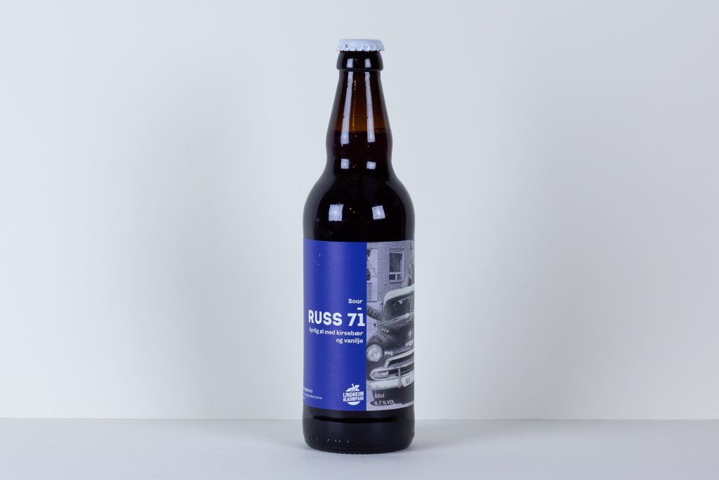 Brown glass beer bottle with a blue label featuring a collage image and the text RUSS 71 on a plain light background