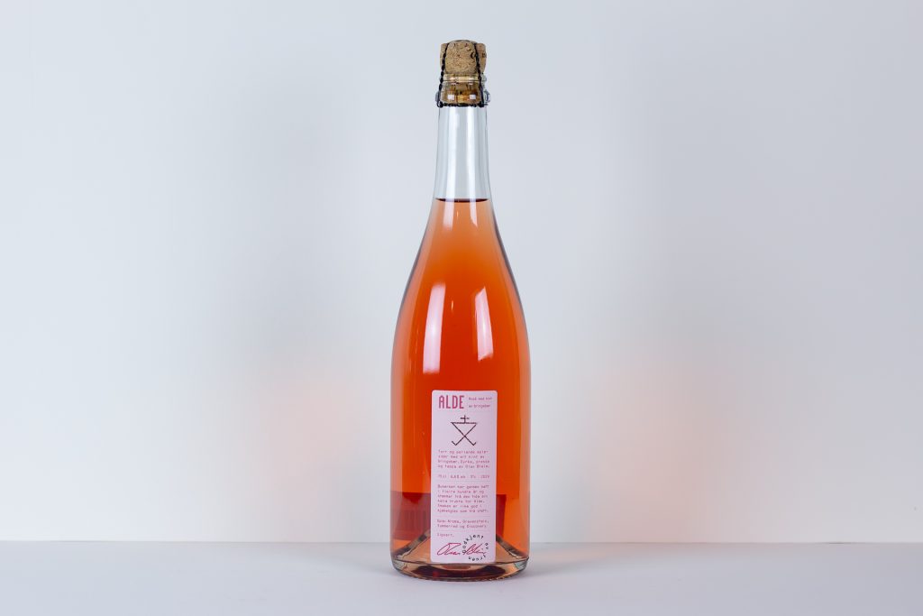 Tall glass bottle of pink sparkling drink with a cork and wire cage label reads ALDE on a white surface