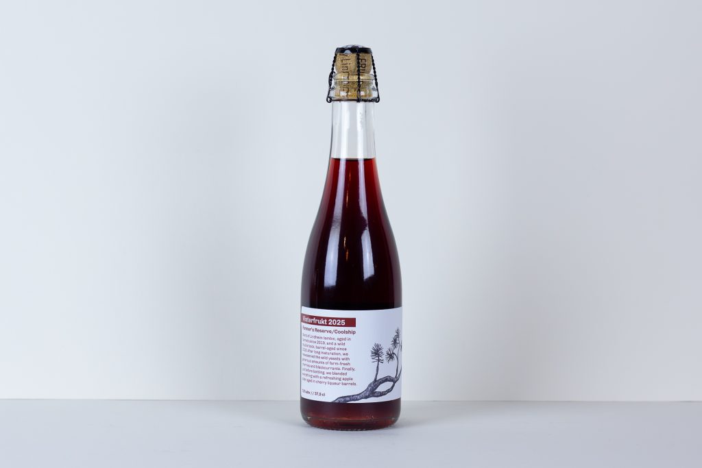 Bottle of dark red liquid with a cork and wire cradle standing on a white surface label visible with Vinterfrukt 2025 text