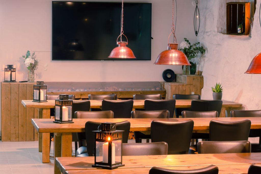 Rustic restaurant dining area with wooden tables and black chairs large red pendant lights lanterns and a wall mounted TV on a white wall