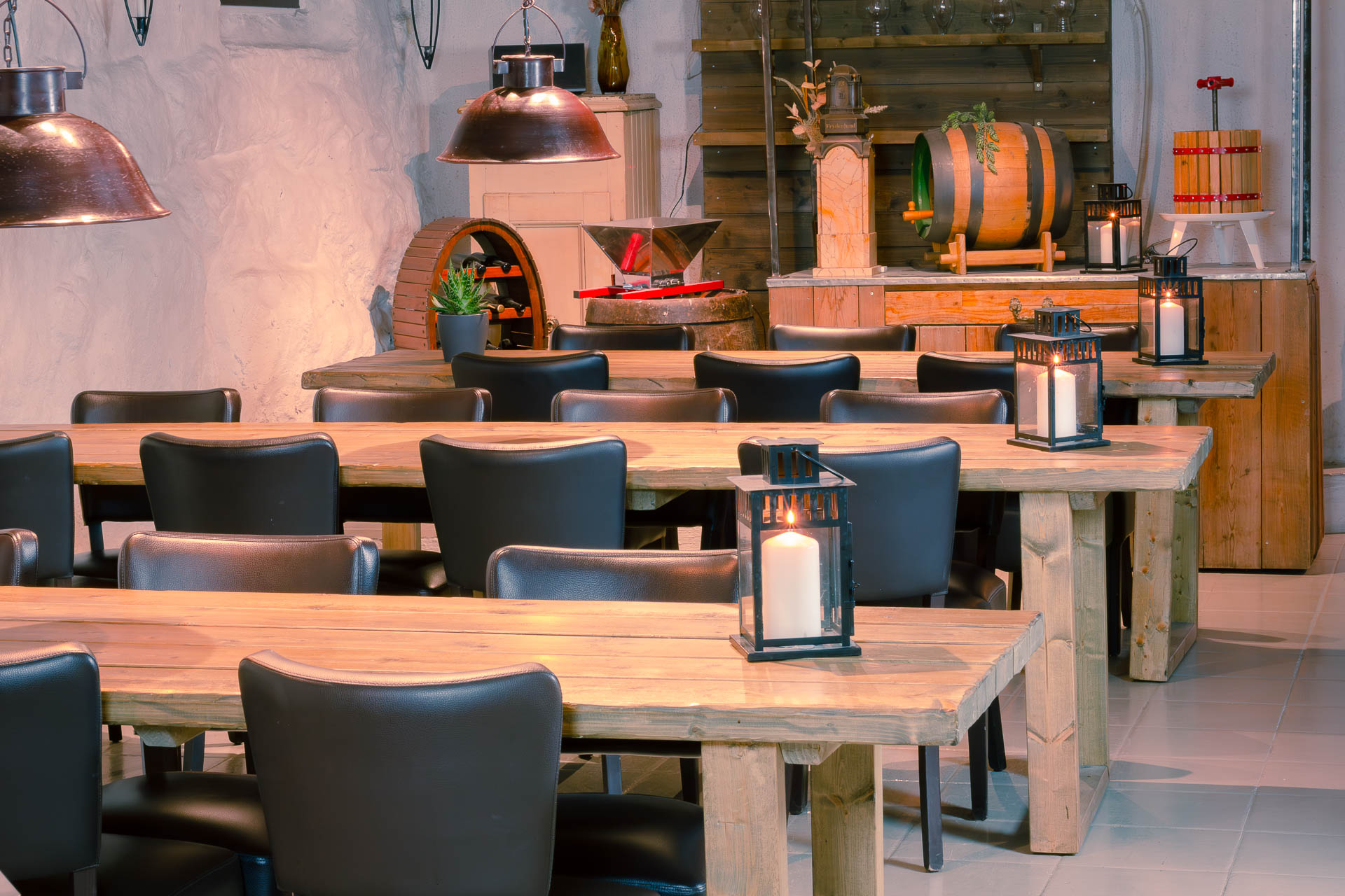 Rustic winery dining area with long wooden tables, black chairs, candle lanterns, and wine barrels in the background.